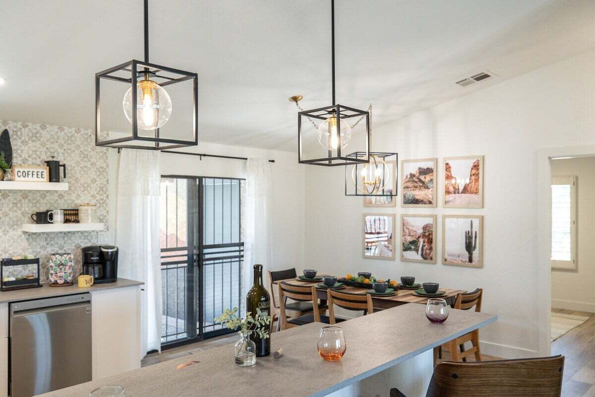 A modern kitchen with a dining area, featuring pendant lights, a coffee station, and framed artwork on the wall.