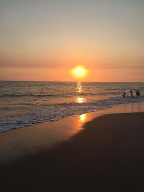 Escena de playa con el sol poniéndose sobre el océano, olas mansas acariciando la orilla y siluetas de personas disfrutando de la vista.