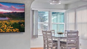 A dining area with a round table, four chairs, a chandelier, a window with blinds, and a painting of a landscape. - Quaint Mid-century Cottage by the Coast (Ventura)