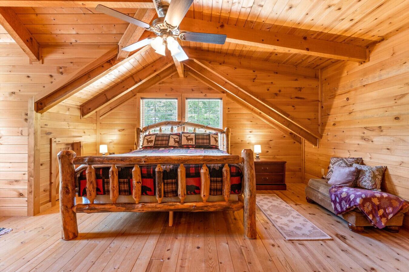 A cozy log cabin bedroom with a wooden bed, a ceiling fan, a nightstand, and a sofa.