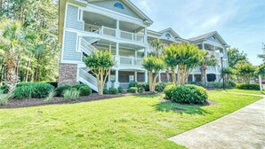 A multi-story residential building with a covered front porch, a central staircase, and a landscaped front yard. - River Crossing 231 (North Myrtle Beach)