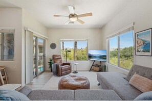 A spacious living room with a ceiling fan, large windows, a comfortable sofa, a leather chair, and a flat-screen TV. - Scottsdale - 12050 N Panorama Dr #208 (Fountain Hills)