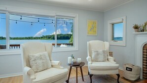 A room with two cream-colored armchairs, a small round table with candles, and a view of a lake through large windows.