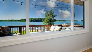 A room with a large window overlooking a lake, a wooden deck, and outdoor seating.