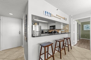 A modern kitchen with a bar area, white cabinetry, and a microwave. There are two wooden stools at the bar. - South Hampton 301 (Myrtle Beach)