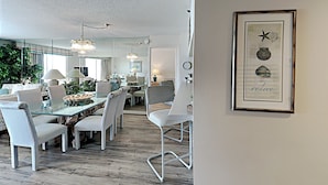 Ein moderner Essbereich mit Glastischen und weißen Stühlen, einer Kristallleuchtre und einem gerahmten Kunstwerk an der Wand.