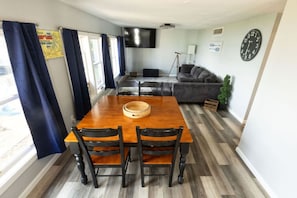 A dining area with a wooden table and chairs, a flat-screen TV mounted on the wall, a sofa, and a clock. - The Jackson Cottage on Grass Lake - Waterfront 2/1 Nautical Cottage With Hot Tub, Firepit & Year-round Fun (Spring Grove)