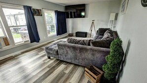 Een woonkamer met een grijze bank, een houten vloer, een statief en een plant in een pot.