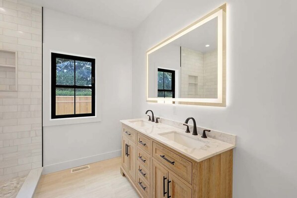 A modern bathroom with a large mirror, two sinks, wooden cabinetry, and a window. - Summit Vista by Rove 4BR Home W/patio & Pool (Westhampton)