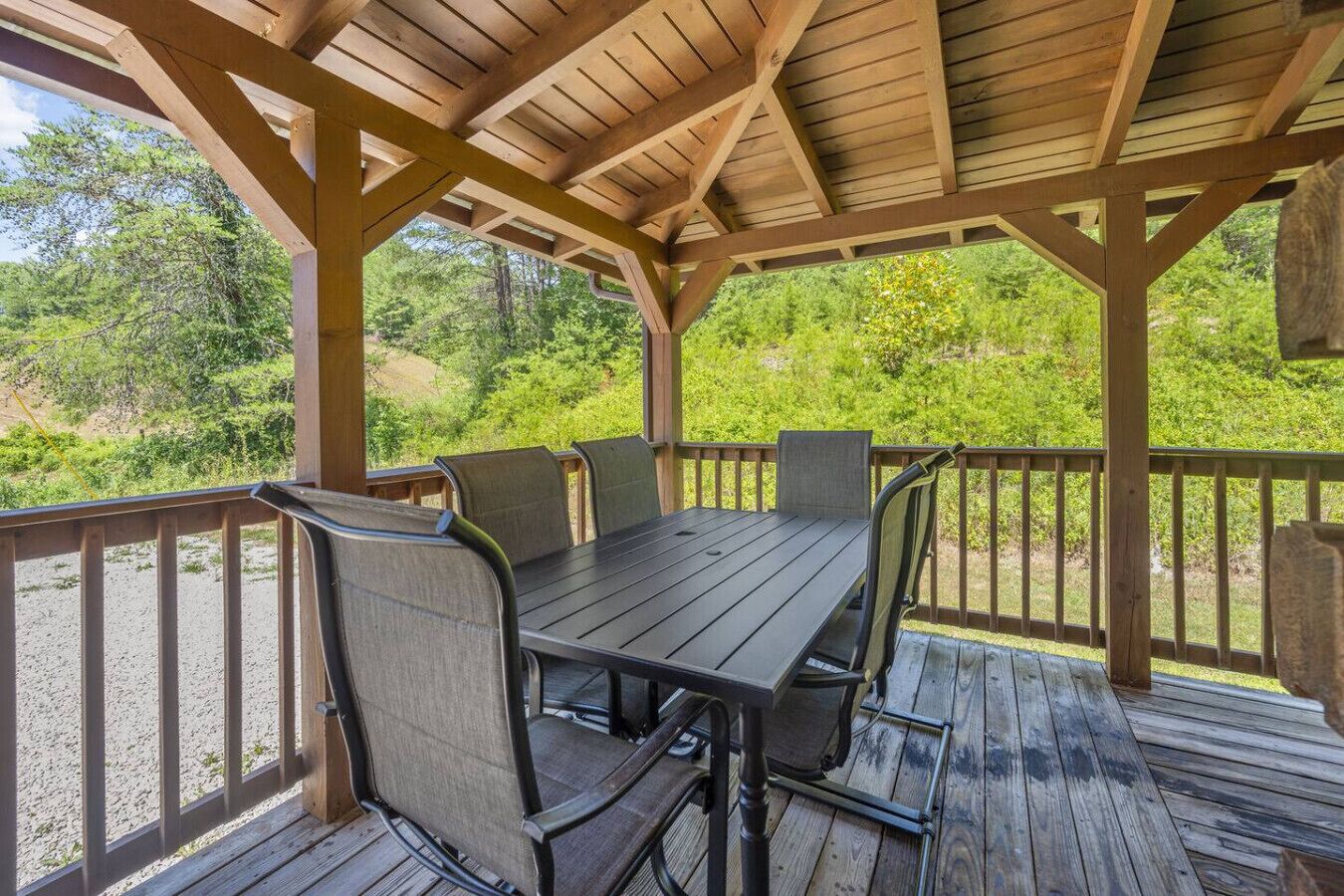 A covered outdoor dining area with a wooden table and chairs, surrounded by a wooden deck and railing, overlooking a forested area.