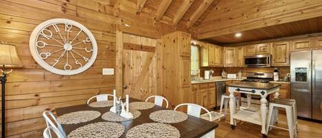 Ein Holzinterieur mit einem für vier Personen gedeckten Esstisch, einer Küchenzeile mit modernen Geräten und einer großen Wanduhr.