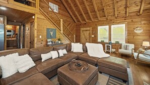 A cozy cabin interior with a wooden ceiling, a sectional sofa, a dining table, and a kitchen area.
