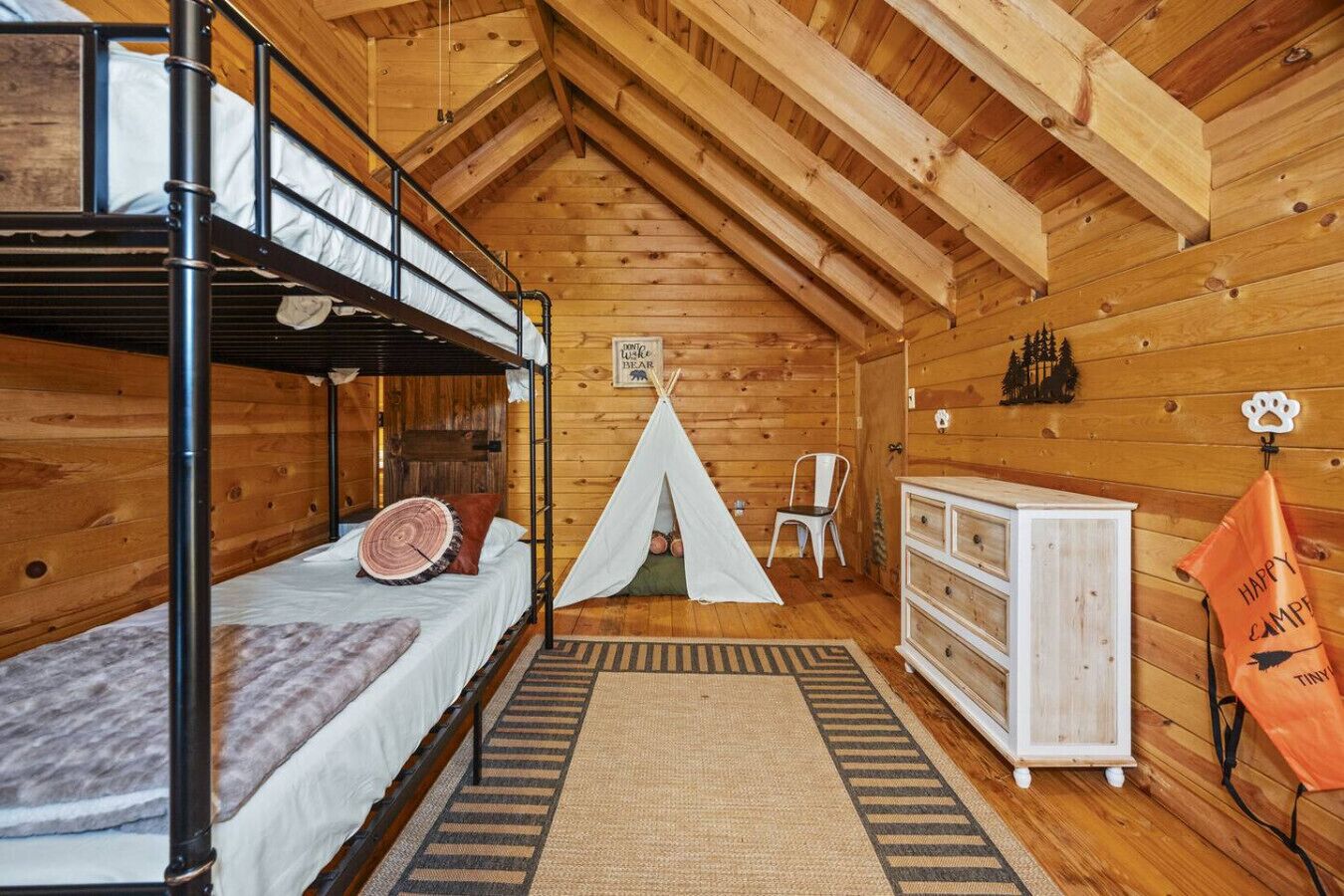 A cozy cabin with a bunk bed, a teepee, a dresser, and a sign that says "Happy Camp Tiny Bear".