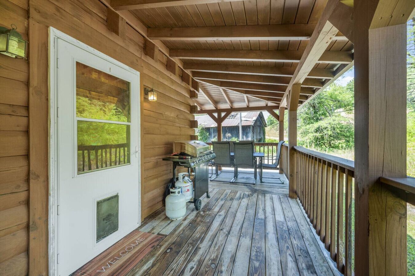 A wooden porch with a grill and seating area, a welcome mat, and propane tanks.