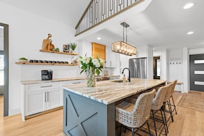 A modern kitchen with a large island, bar stools, a hanging light fixture, and a stainless steel refrigerator. - Niblick on Norman (Denver)