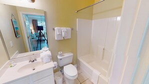 Ein Badezimmer mit Badewanne, Toilette und Waschbecken. Über dem Waschbecken befindet sich ein Spiegel, und Handtücher hängen an der Wand.