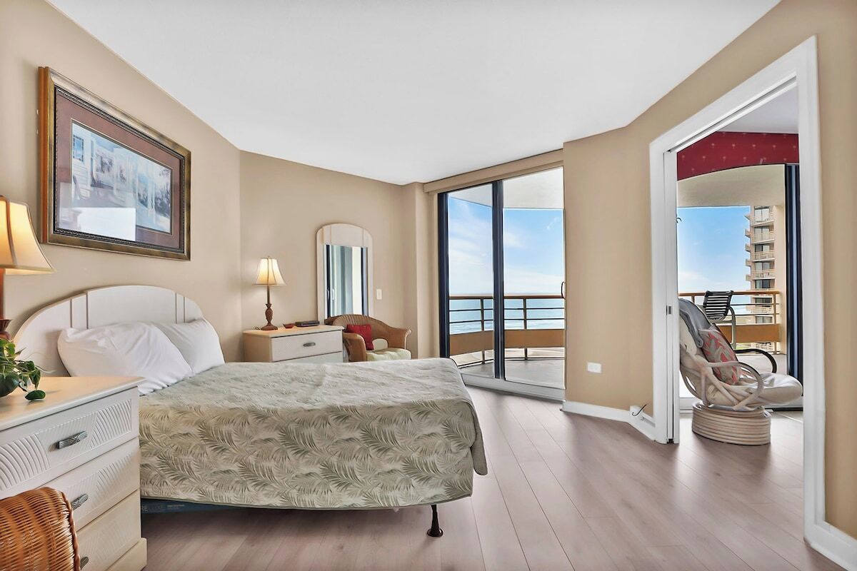 A spacious bedroom with a large bed, a bedside table with a lamp, a dresser, and a framed picture on the wall. There is a balcony with a chair and a view of the sea.