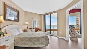 Une chambre spacieuse avec un grand lit, une table de chevet avec une lampe, une commode et un tableau encadré au mur. Il y a un balcon avec une chaise et une vue sur la mer.