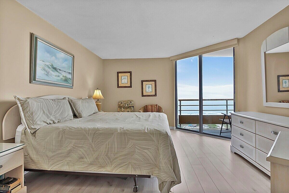 A bedroom with a bed, a dresser, a nightstand, and a large window offering a view of the ocean.