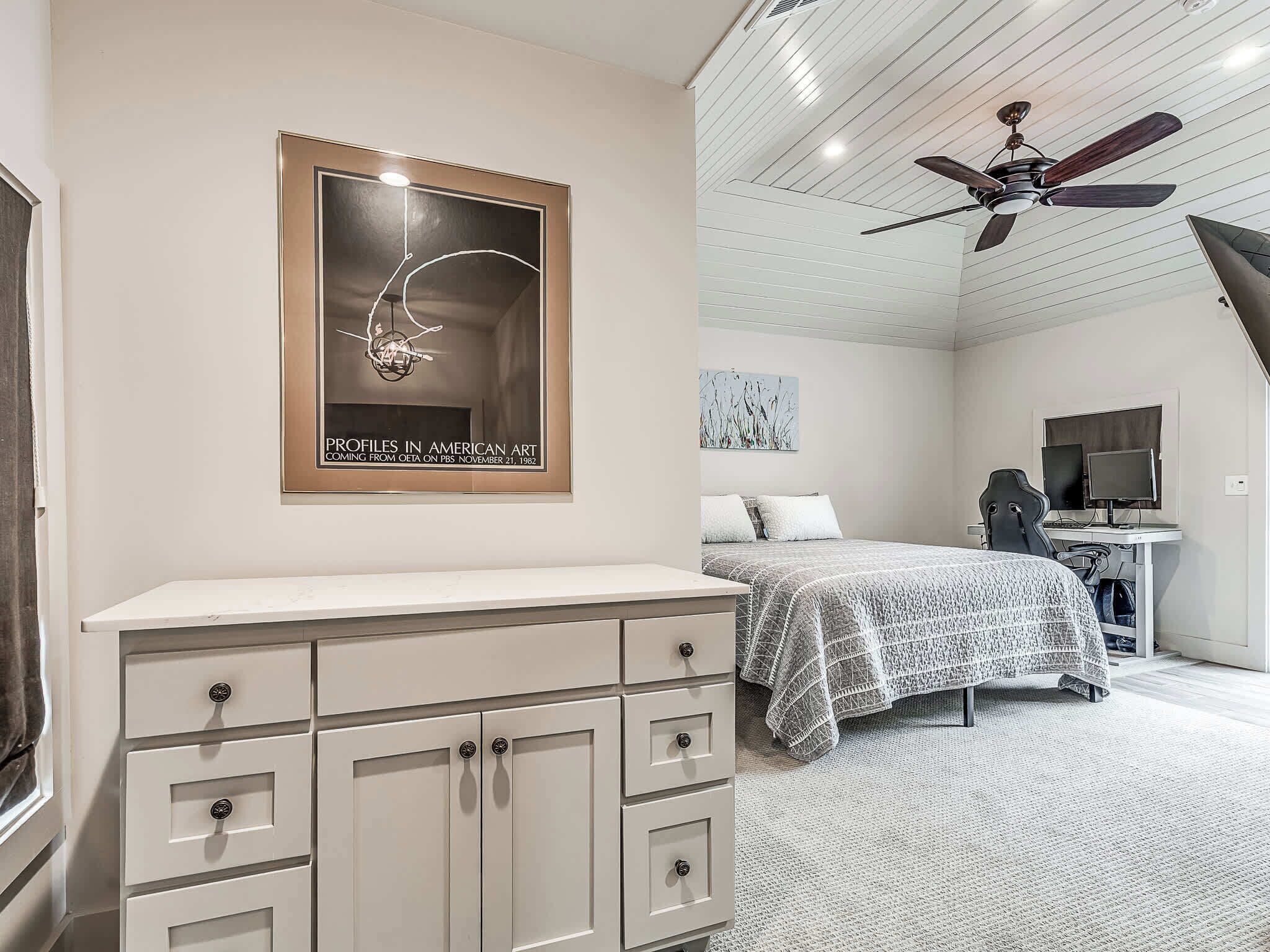 A bedroom with a bed, a desk with a computer, a ceiling fan, and a framed artwork on the wall.