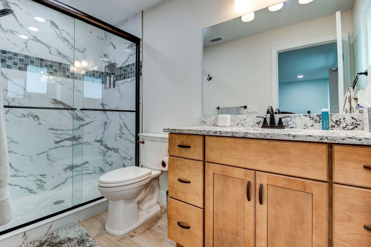 A bathroom with a glass shower enclosure, a toilet, a marble countertop, and wooden cabinets.