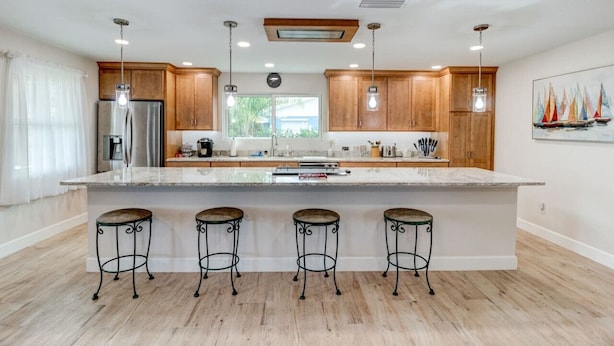 A modern kitchen with wooden cabinets, a stainless steel refrigerator, a large island with stools, pendant lights, and a window with a view of greenery. - Cape Key Updated Beach Bungalow (Ponce Inlet)
