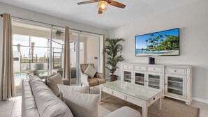 Ein Wohnzimmer mit einer Couch, einem Glastisch, einem Fernseher und Blick ins Freie.