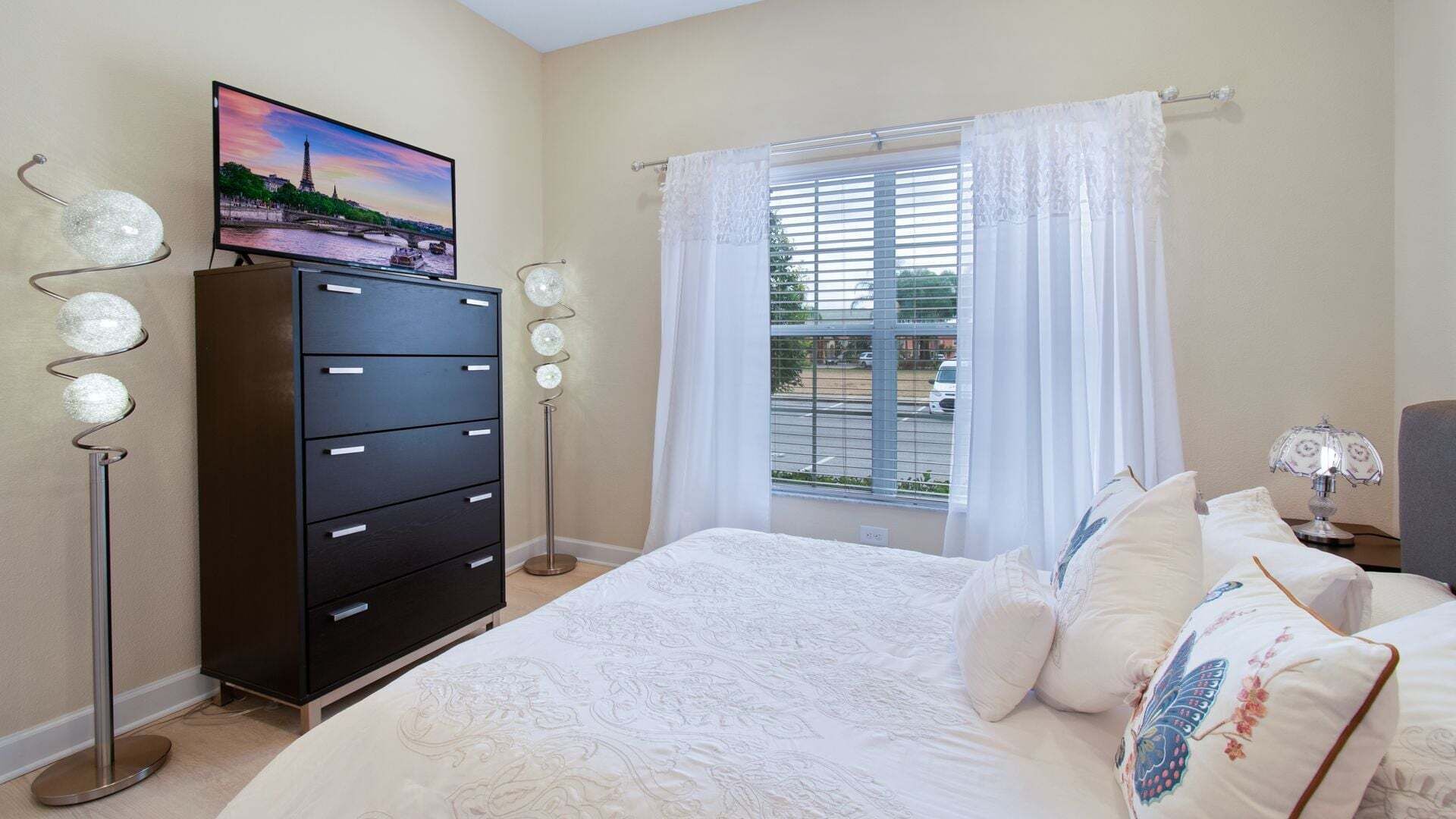 Une chambre à coucher avec un lit, une commode, un téléviseur et une fenêtre avec des rideaux.