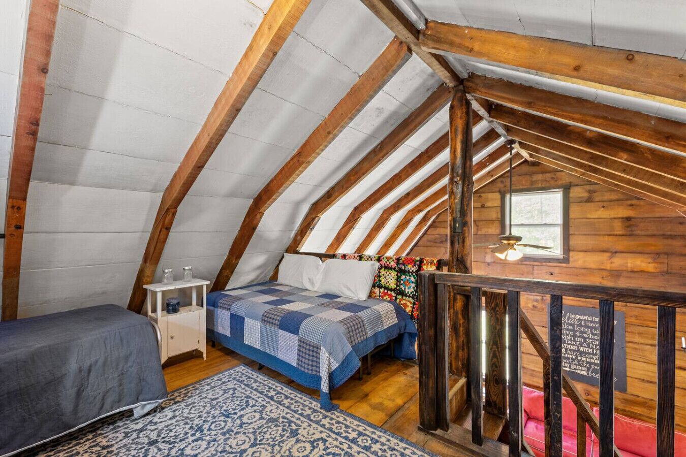 Une chambre mansardée chaleureuse, avec un plafond en pente, des poutres apparentes, un lit recouvert d’une courtepointe, une petite table avec une lampe et une fenêtre offrant une belle vue.