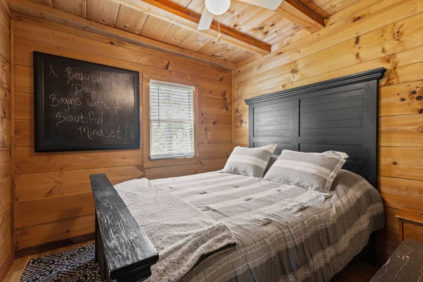 A wooden cabin bedroom with a bed, a chalkboard with a motivational quote, and a window with blinds.