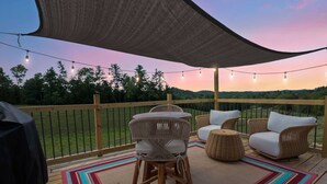 An outdoor deck with wicker furniture, a colorful rug, and a string of lights against a sunset backdrop. - Wifi, Firepit, Fishing Pond - Farmview (Hazel Green)