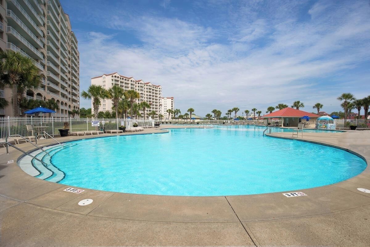 A large outdoor swimming pool with a curved design, surrounded by a white fence and palm trees.