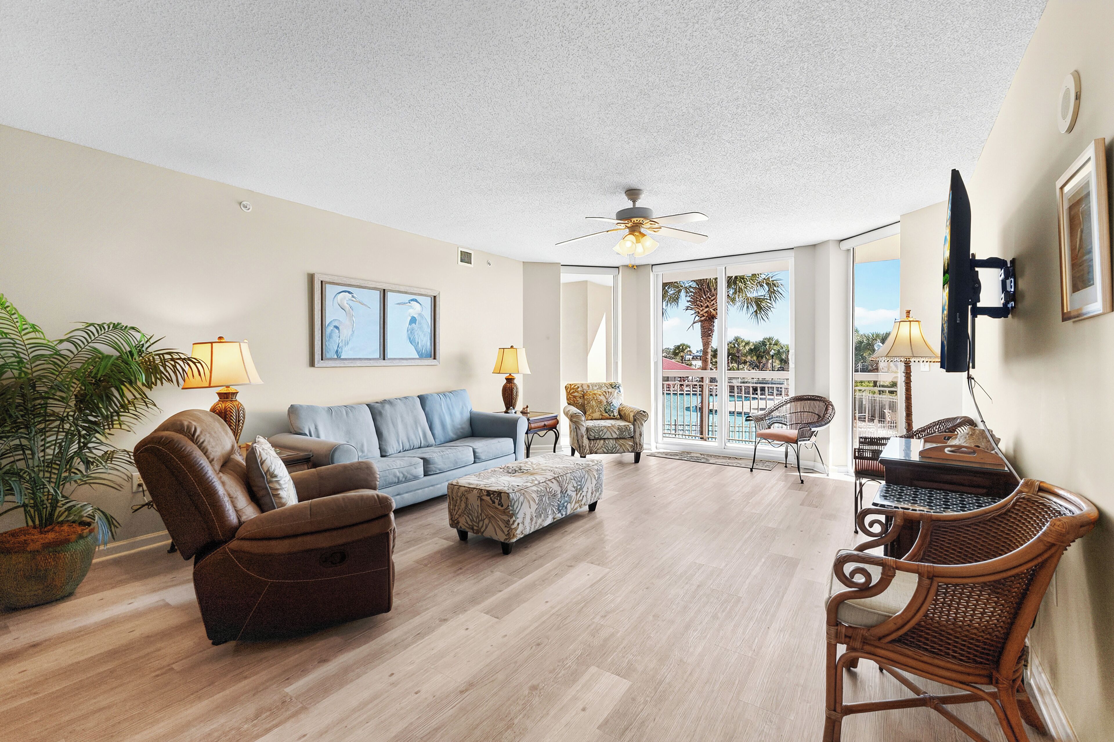 A spacious living room with a blue sofa, brown recliner, and ottoman, a ceiling fan, and a large window offering a view of palm trees.