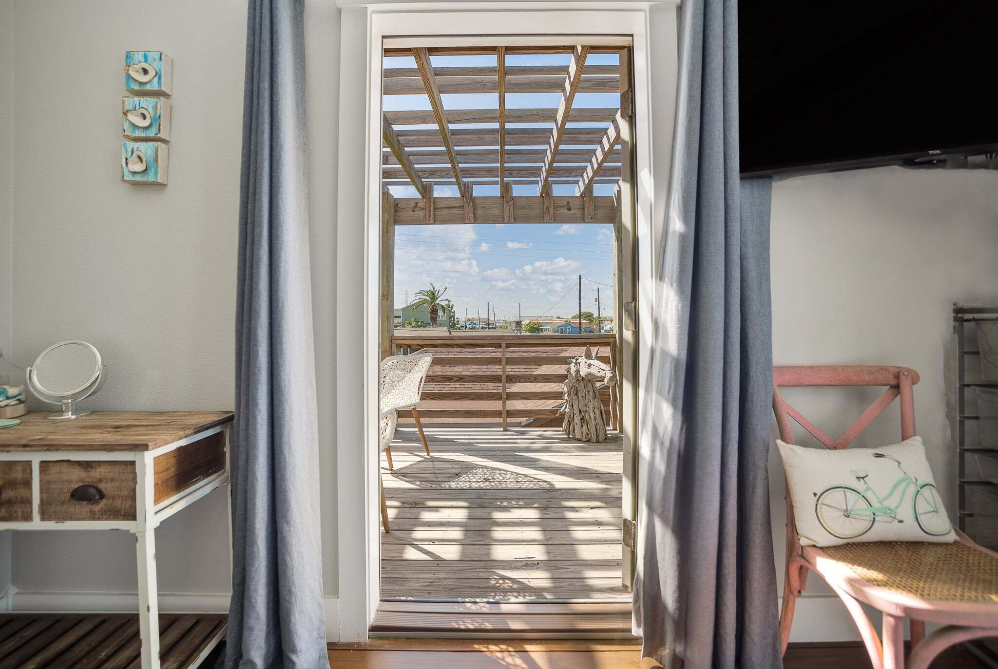 Et værelse med et træskrivebord, en stol, et lille bord med et spejl, et vindue med gardiner og udsigt til en balkon med en træpergola.