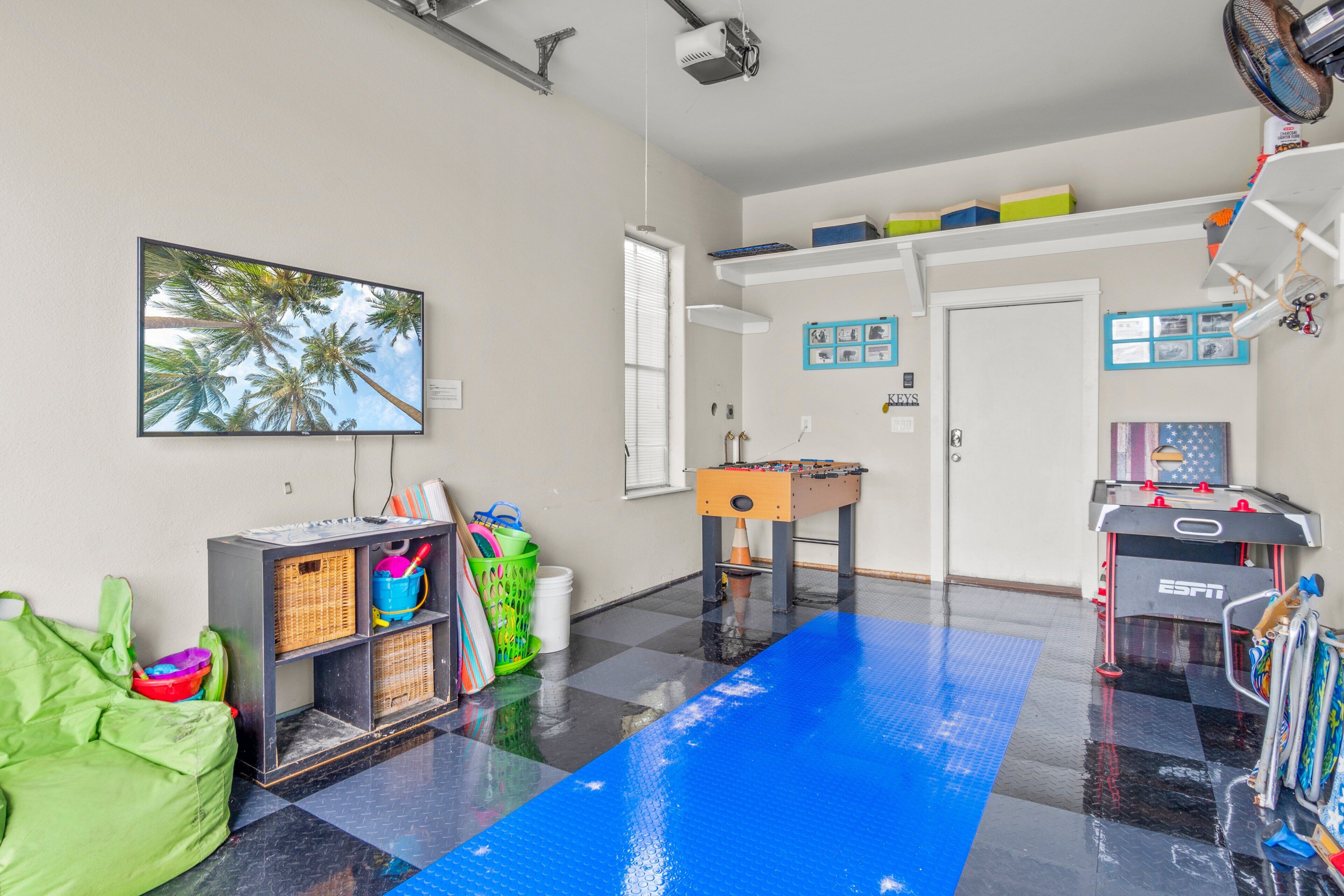 A room with a blue mat, a foosball table, a shelf with toys, and a window with blinds.