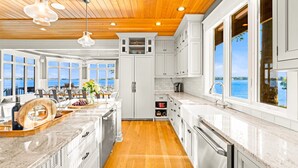 Een moderne keuken met witte kasten, een houten plafond en grote ramen met uitzicht op het water.