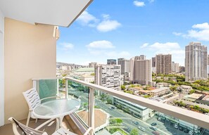 A balcony with a glass table and chairs, overlooking a cityscape with high-rise buildings and a road. - Stunning Ala Moana 2501 (Honolulu)