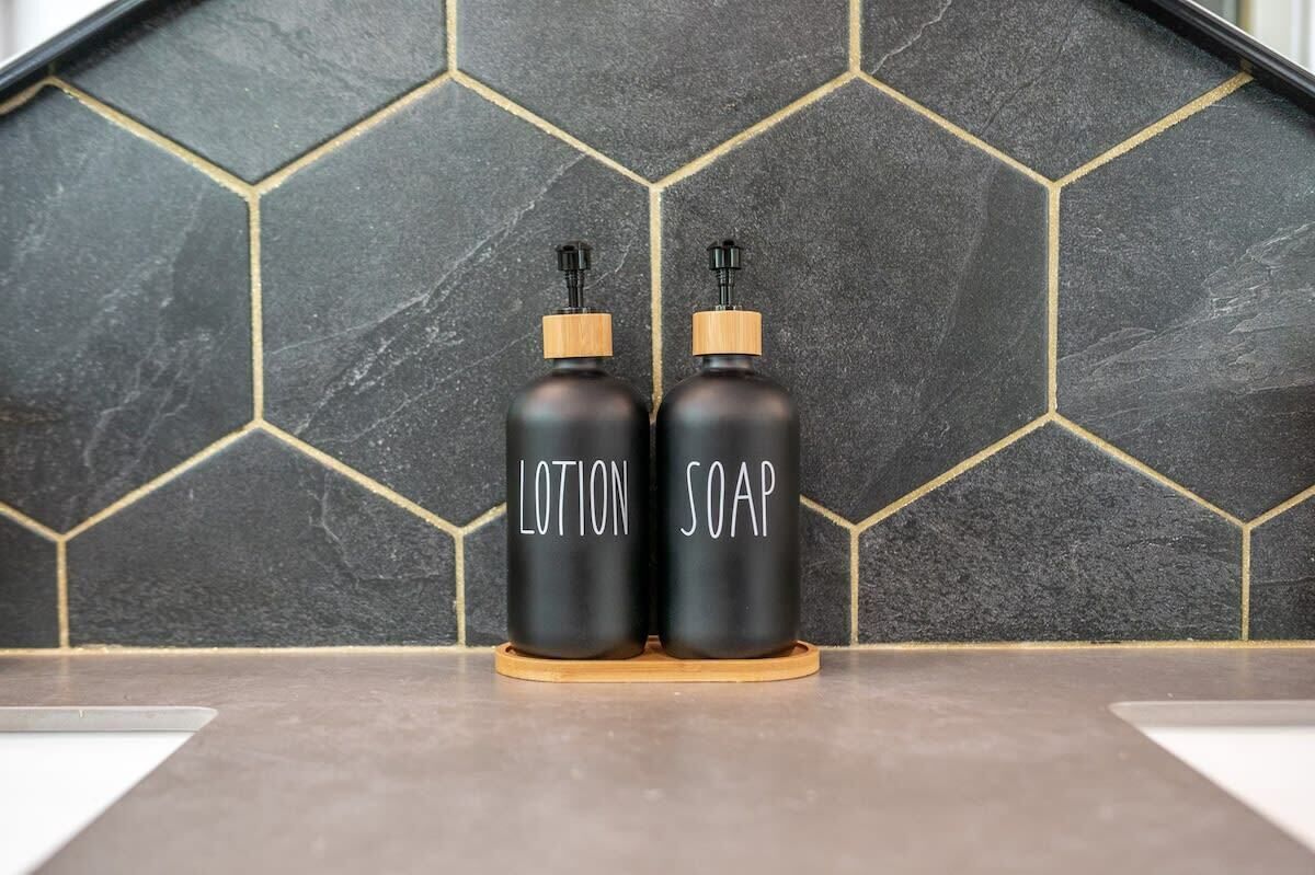 Deux flacons noirs étiquetés « LOTION » et « SAVON » sont posés sur un sous-verre en bois, devant un mur recouvert de carreaux hexagonaux.