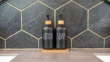 Deux flacons noirs étiquetés « LOTION » et « SAVON » sont posés sur un sous-verre en bois, devant un mur recouvert de carreaux hexagonaux.