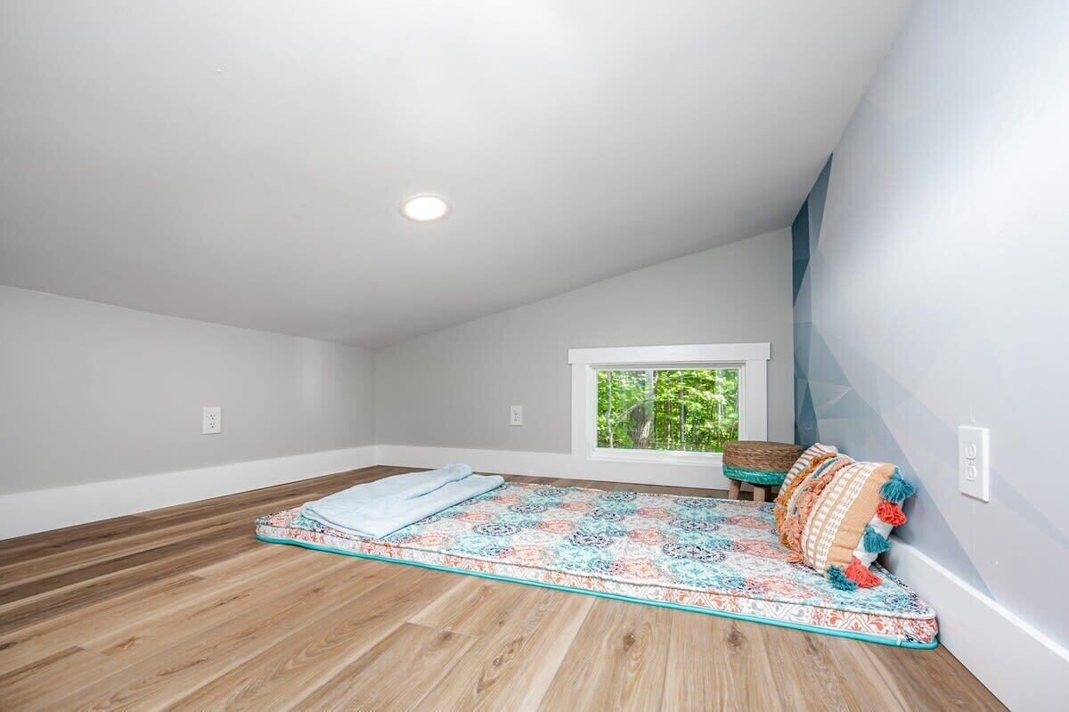 An attic bedroom with a wooden floor, a colorful quilt on the bed, a window with a view of greenery, and a small table with a basket.