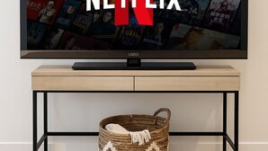 Television ruudulla näkyy Netflixin käyttöliittymä, ja siinä on esillä useita elokuvajulisteita. Televisio on kiinnitetty seinälle, ja sen alla on puinen konsolipöytä, jonka alla on punottu kori.