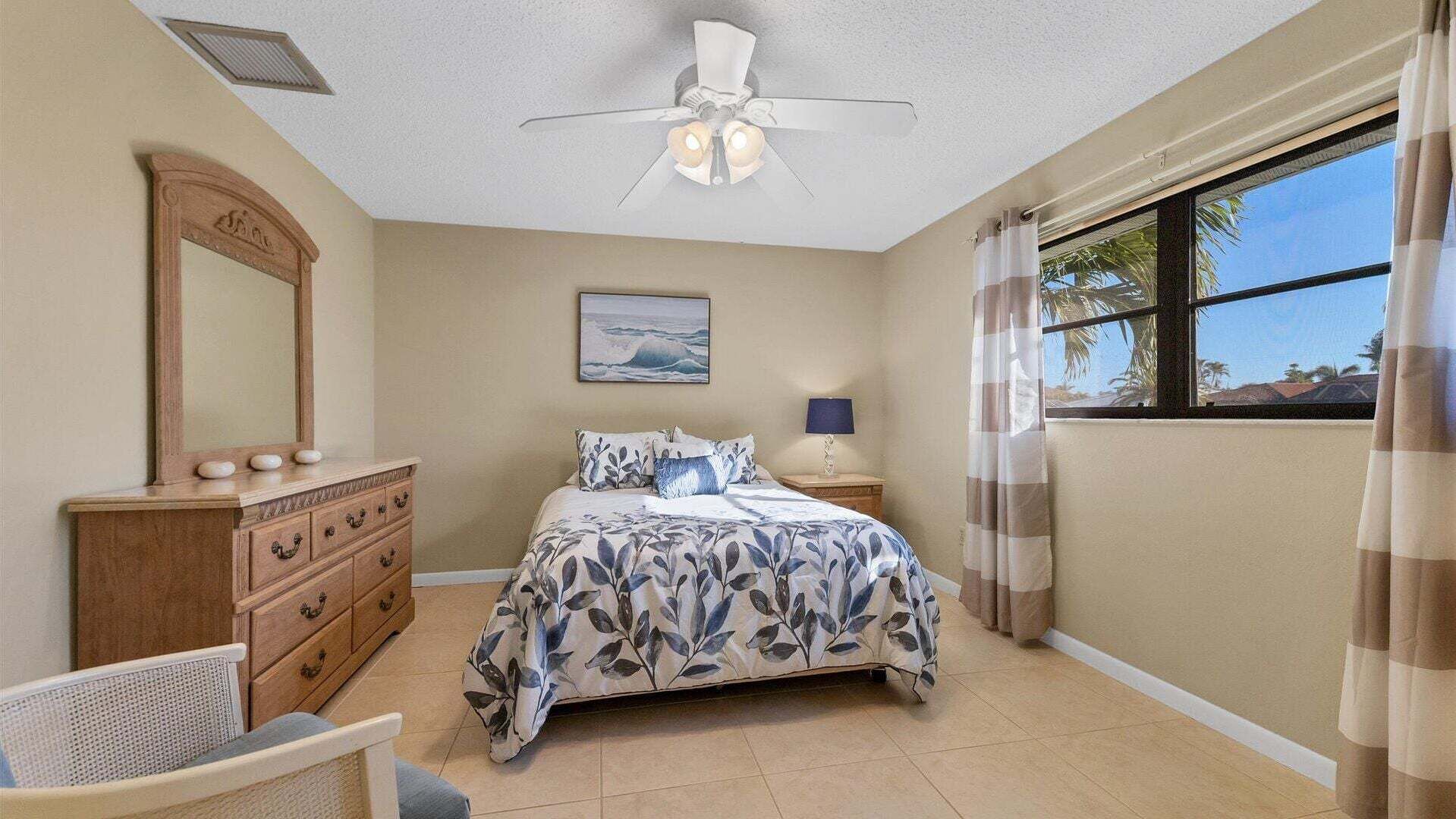 A bedroom with a bed, dresser, chair, ceiling fan, window, and artwork.