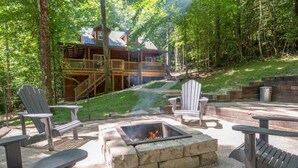 An outdoor seating area with Adirondack chairs, a fire pit, and a view of a cabin in a wooded area. - Wifi & Hot Tub, Large Family - Climb On in Rrg! (Rogers)