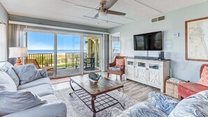 En rummelig stue med sofa, sofabord og fjernsyn. Der er en balkon med udsigt over havet.