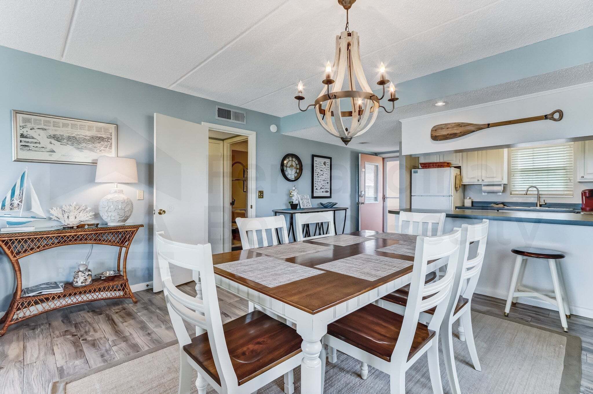 A dining area with a wooden table, white chairs, a chandelier, and a kitchen in the background.
