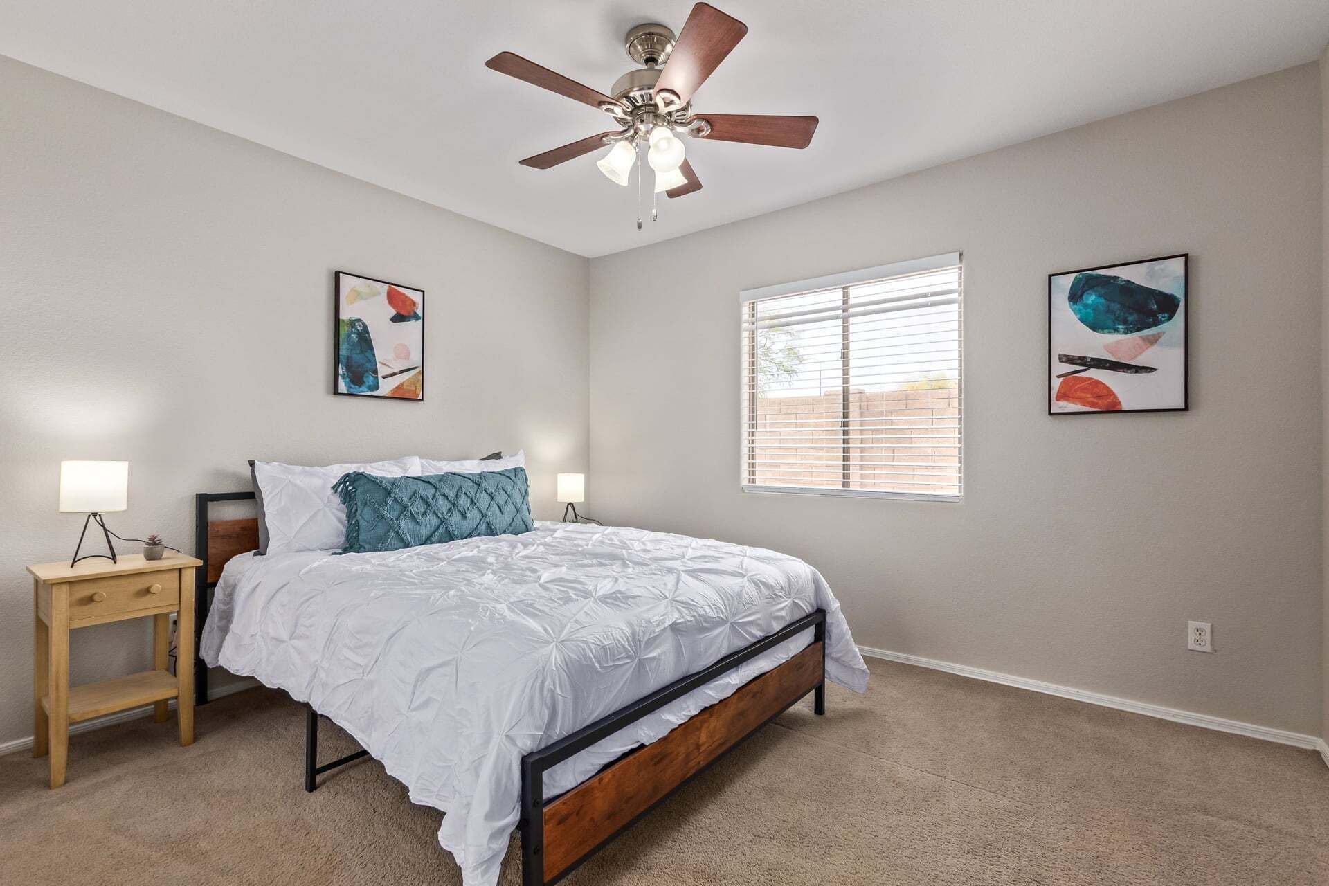 A bedroom with a bed, a nightstand, a ceiling fan, two wall-mounted artworks, and a window with blinds.