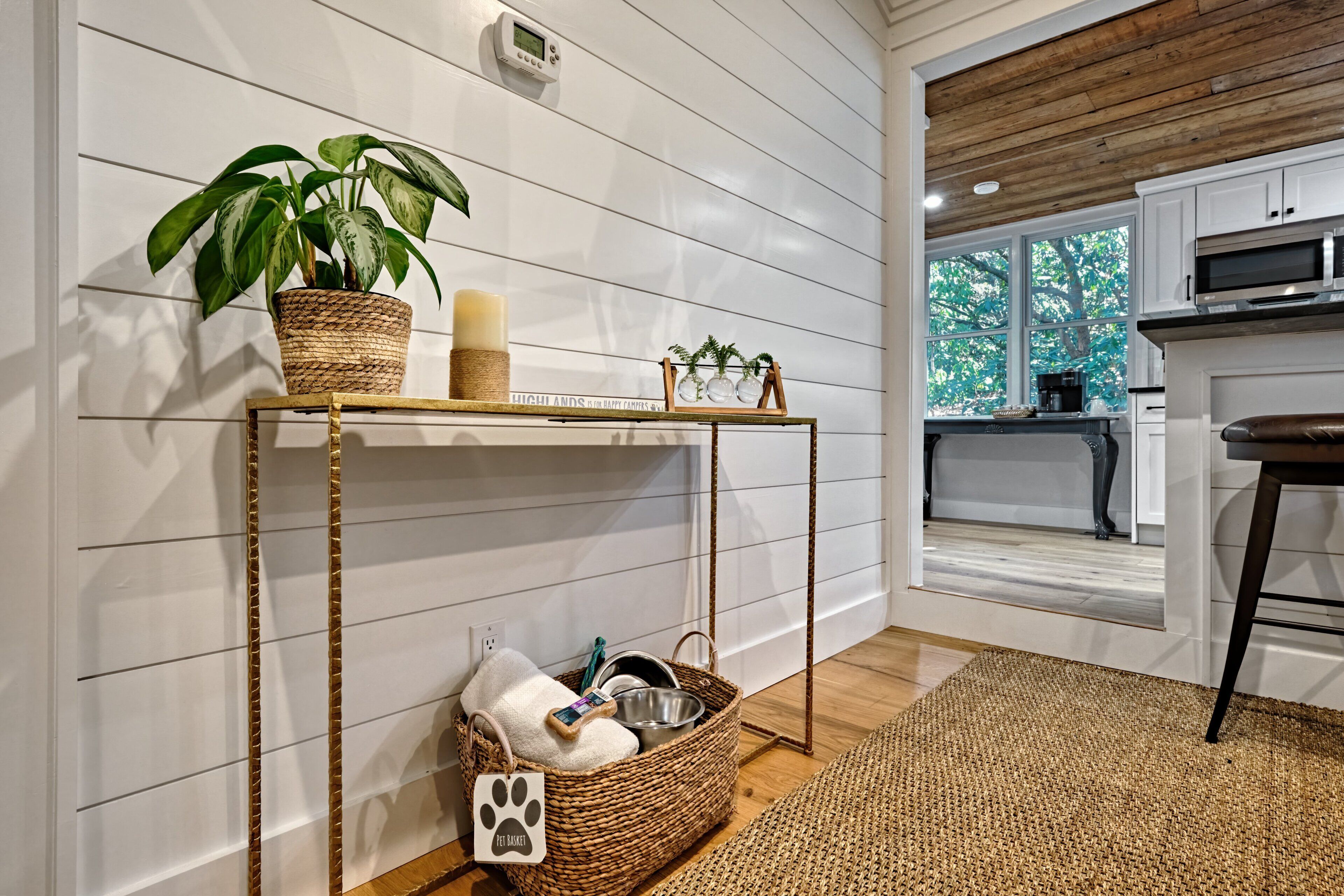 A room with a wooden floor, white walls, a kitchen area with a window, and a wicker table with a plant and candle.
