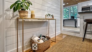 Una habitación con piso de madera, paredes blancas, una zona de cocina con ventanal y una mesa de mimbre con planta y vela.