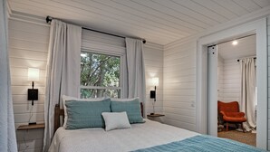 Un dormitorio con cama, dos lámparas de noche, una ventana con cortinas y una silla en la habitación contigua.
