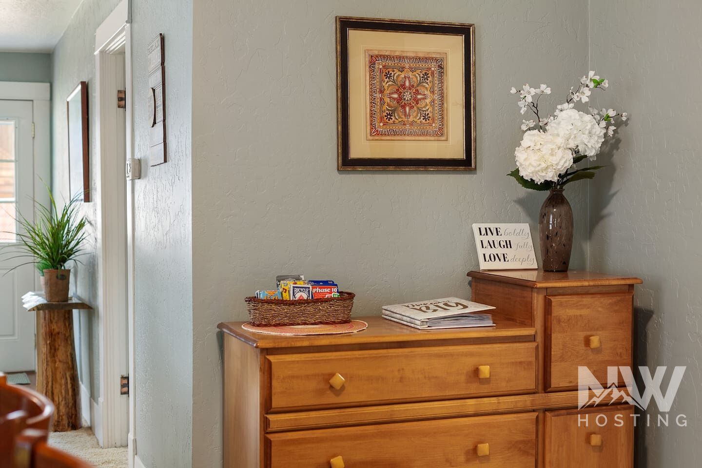 A room with a framed artwork, a vase with white flowers, a basket with items, and a sign with motivational words.